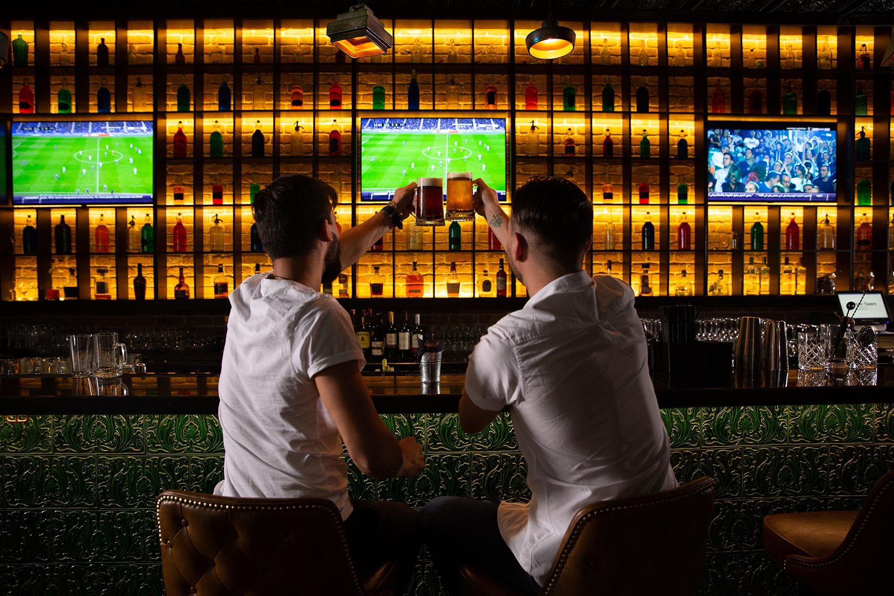 Homens torcendo em bar com jogo de futebol no fundo